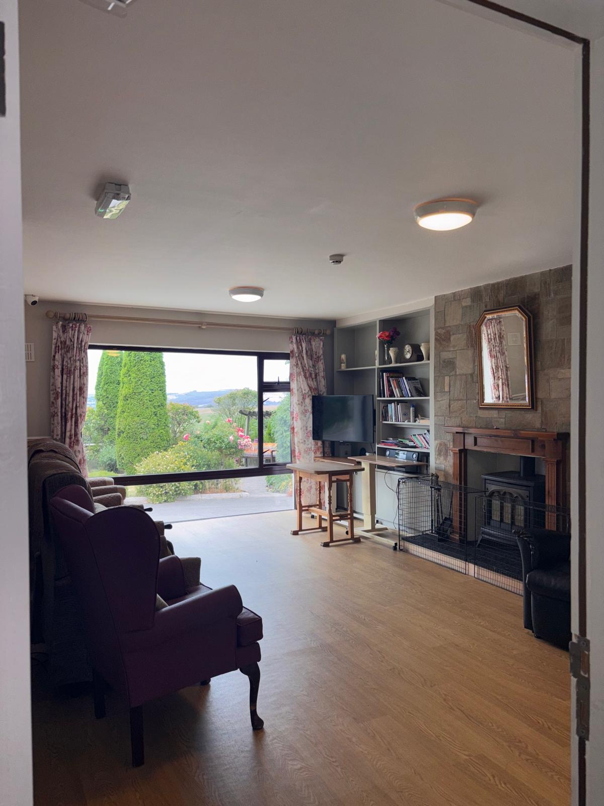 The residents' sitting room at Norwood Grange with armchairs, a wide window onto the garden and a stone fireplace